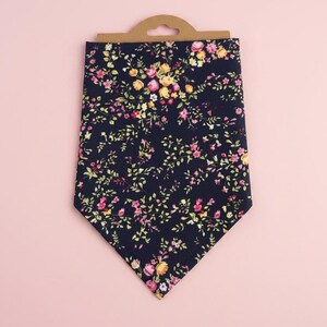 May include: A navy blue floral patterned necktie with a brown cardboard hanger. The tie features a repeating pattern of pink and yellow flowers with green leaves. The background is a solid light pink.