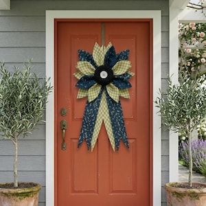 May include: A fabric flower wreath featuring alternating petals of navy blue floral print and light green gingham check fabric. The center is a black circular piece with a white four-hole button. Two long fabric tails hang down from the center.