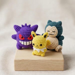 Può includere: Tre figure di Pokémon all'uncinetto: un Gengar viola con occhi rossi e un ampio sorriso, un Pikachu giallo con un colletto bianco e un Snorlax blu e beige. Le figure sono esposte su un blocco di legno chiaro.