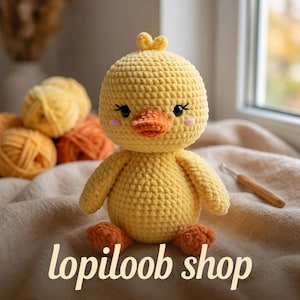 May include: A handmade yellow crochet duckling with an orange beak and feet. The duckling has black eyelashes and pink cheeks. The text "lopiloob shop" is displayed at the bottom. Yarn balls are in the background.