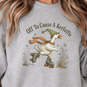 May include: A sport grey crewneck sweatshirt featuring a graphic of a white goose wearing a green patterned hat and an orange scarf while roller skating. The text above the goose reads Off To Cause A Kerfuffle in dark green lettering.