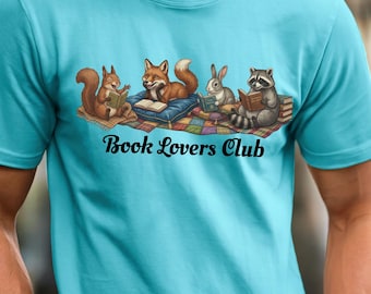 May include: A bright turquoise short-sleeve t-shirt featuring a graphic of a squirrel, fox, rabbit, and raccoon reading books on a colorful patchwork quilt. The text "Book Lovers Club" is printed in black script lettering below the animals.