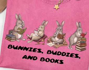 May include: A pink short-sleeved t-shirt featuring a graphic of four rabbits reading books. The text below the rabbits reads "BUNNIES, BUDDIES, AND BOOKS". The shirt has a Comfort Colors label at the neck, and a gold-toned star pendant necklace is placed on top.