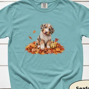 May include: A seafoam green short-sleeve t-shirt featuring a graphic of a brown and white puppy sitting in a pile of orange and brown autumn leaves next to a small striped pumpkin. The shirt has a Comfort Colors brand tag at the inner neckline.