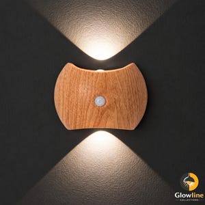 May include: A wooden wall sconce with a light brown finish and a motion sensor. The sconce has a curved, symmetrical design and emits light upwards and downwards, creating a unique lighting effect. The sconce is mounted on a dark gray wall.
