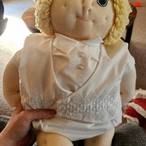 May include: A vintage doll with a soft, fabric body and a smiling face. The doll has blue eyes, a small pink mouth, and blonde yarn hair. It is dressed in a white outfit with a decorative embroidered vest.
