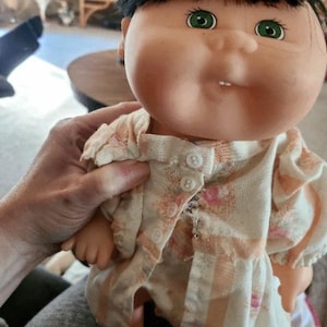 Może przedstawiać: Zabytkowa lalka Cabbage Patch Kid z czarnymi włosami i zielonymi oczami. Lalka ma na sobie brzoskwiniowo-białą sukienkę w kwiaty z detalami guzików. Lalka jest trzymana przez osobę.