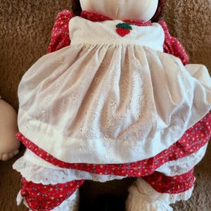 Può includere: Bambola di pezza fatta a mano con un viso sorridente, capelli di filo marrone e occhi ricamati blu. La bambola indossa un vestito bianco con un'applicazione di fragola e rifiniture floreali rosse. Le gambe della bambola sono coperte di tessuto floreale rosso e scarpe all'uncinetto bianche.