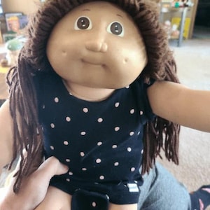 May include: A Cabbage Patch Kids doll with brown yarn hair and a round face. The doll is wearing a navy blue onesie with pink polka dots. The doll is being held by a person.