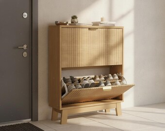 May include: A light wood shoe cabinet featuring a fluted front panel design, a drop-down storage compartment with internal shelving, and four sturdy legs. The cabinet holds multiple pairs of sneakers and casual shoes on its open shelves.