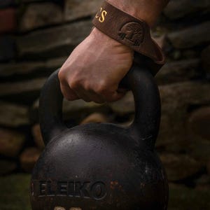 Peut inclure: Une sangle de levage en cuir marron foncé avec un logo en relief représentant un arbre et une barre, portée au poignet tout en tenant une kettlebell en fonte noire. La kettlebell porte le texte ELEIKO et le chiffre 32 en relief.