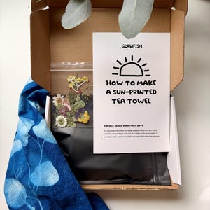 May include: A craft kit in a cardboard box containing a white instruction card titled HOW TO MAKE A SUN-PRINTED TEA TOWEL by GLOWFISH, a clear plastic bag of dried flowers, and a blue tea towel with a white leaf print pattern on the side.