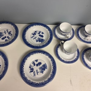 May include: A collection of white and blue porcelain dishware, including plates, saucers, and teacups. The plates and saucers have a blue border and a central blue illustration. The teacups are white with blue accents. The set is arranged on a white surface.