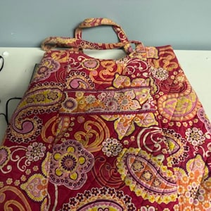 Puede incluir: Un bolso tote acolchado con un vibrante estampado de cachemira en tonos de rojo, rosa, amarillo y naranja. El bolso cuenta con un asa superior y un diseño grande y abierto. La tela tiene una textura acolchada, y el patrón incluye motivos florales y de remolino.
