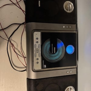 May include: A Philips micro music system with a central silver and black console featuring a circular CD player, a blue digital display, and various control buttons. Two black speakers with silver circular accents are attached to the sides.