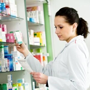Puede incluir: Un farmacéutico con bata blanca de laboratorio está frente a estantes llenos de varias cajas y frascos de medicamentos. El farmacéutico sostiene una pequeña caja rectangular blanca de medicamentos en una mano y un documento de papel.
