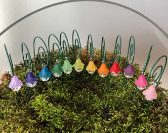 May include: A display of colorful miniature fairy lights in a terrarium setting. The lights are arranged on green wire stems, each topped with a translucent bell-shaped shade in various colors. The lights are set in a bed of green moss.