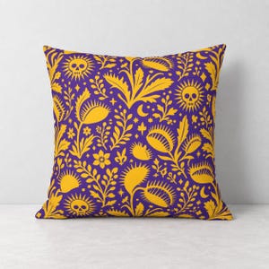Puede incluir: Un cojín cuadrado con un fondo morado vibrante y un patrón repetitivo de ilustraciones amarillas. El diseño incluye plantas carnívoras atrapamoscas, soles con centros de calavera, lunas crecientes, estrellas pequeñas y diversas ramas botánicas.