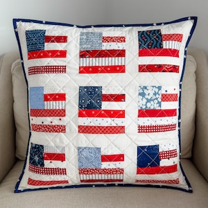 Puede incluir: Cojín decorativo cuadrado con un diseño de patchwork de banderas americanas. Cada bandera está compuesta por rayas rojas y blancas y un campo azul con estrellas u otros patrones. El cojín tiene un fondo blanco, un borde azul y un patrón acolchado.