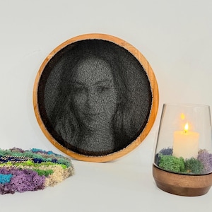 Può includere: Un ritratto rotondo di string art con cornice in legno. Sono presenti anche una candela accesa in un portacandele di vetro e una colorata composizione di muschio. Il ritratto è realizzato con filo nero, creando un'immagine dettagliata. La candela è bianca e il muschio è un mix di verde, viola e beige.