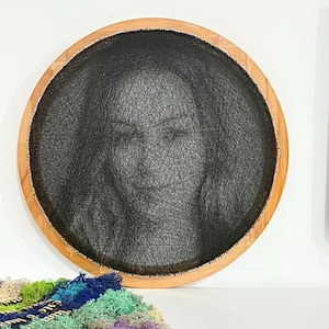 May include: Round wall art featuring a portrait created with black thread on a dark background, framed by a natural wood border. The artwork depicts a face with closed eyes. Decorative moss in various colors is visible below.