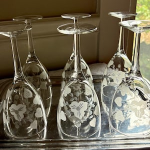 May include: A set of six clear glass wine glasses arranged upside down on a silver tray. Each glass features a delicate etched floral pattern with leaves and blossoms on the bowl. The stems are slender and clear, leading to circular bases.