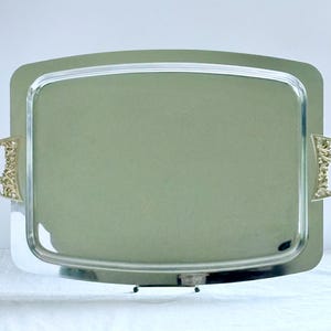 May include: A rectangular silver-toned serving tray with a polished, reflective surface and rounded corners. The tray features two ornate, gold-toned metal handles on the left and right sides, each decorated with intricate, raised floral scrollwork.