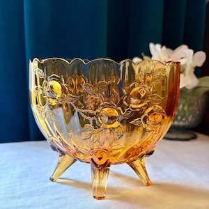 May include: Amber-colored, footed glass bowl with a floral rose pattern. The bowl has a scalloped rim and three delicate legs. The embossed roses and leaves add a vintage touch to this decorative piece. A beautiful accent for any home.