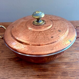 May include: A round, hammered copper casserole dish with a matching lid. The pot features two curved brass side handles and a central brass knob on the lid. The copper surface displays a warm, metallic finish with visible signs of natural patina.
