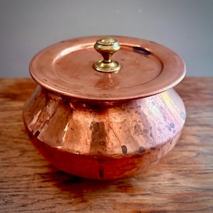 May include: A round copper pot with a matching lid and a small brass knob handle. The copper surface features a warm, metallic finish with visible natural patina and slight variations in color, giving it a textured, aged appearance.