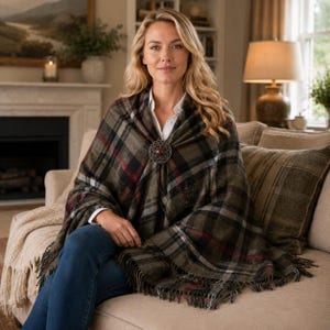 May include: A plaid shawl in shades of olive green, black, and red, featuring a circular metal brooch with a central brown stone at the closure. The shawl has a fringed hem and is worn over a white collared shirt and blue denim jeans.