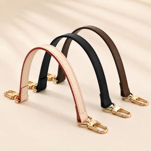 May include: Three arched bag handles in cream, black, and brown, each featuring gold-toned metal lobster clasps at both ends. The cream handle has contrasting red edges, while all three straps are constructed with visible stitching along the sides.