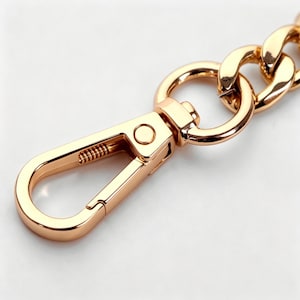May include: A polished gold-tone metal lobster claw clasp attached to a circular jump ring, which connects to a section of a thick curb chain. The hardware features a smooth, reflective finish and a spring-loaded mechanism for secure attachment.