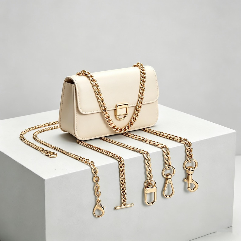 May include: A cream-colored rectangular shoulder bag featuring a gold-tone metal chain strap. The bag has a front flap with a gold-tone rectangular clasp. Several additional gold-tone chain segments with various metal clips and fasteners are attached.
