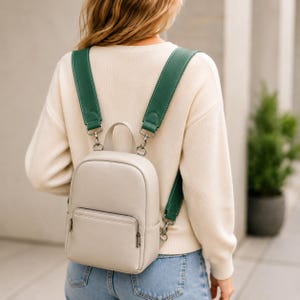 May include: A small, light beige backpack featuring a smooth, solid-colored exterior, a front zippered pocket, and a top handle. The bag is equipped with wide, detachable dark green shoulder straps that attach with silver-toned metal hardware.
