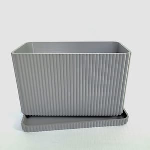 May include: A gray rectangular planter with vertical ribbed detailing. The planter sits on a matching tray. The design is modern and minimalist, suitable for indoor or outdoor use. The planter is made of a durable material.