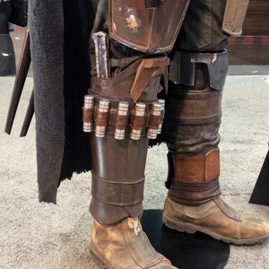 May include: Detailed view of a costume's lower leg armor, featuring brown leather boots and leg coverings. The armor includes a series of cylindrical metallic cartridges and a holster for a weapon. The costume has a weathered, battle-worn appearance.