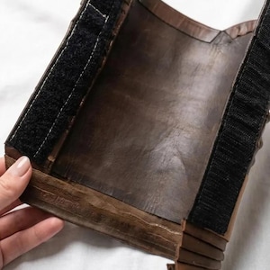 May include: A brown leather wrist cuff with black hook-and-loop fasteners. The cuff is made of layered leather and has a dark, aged appearance. The cuff is held by a hand against a white background.