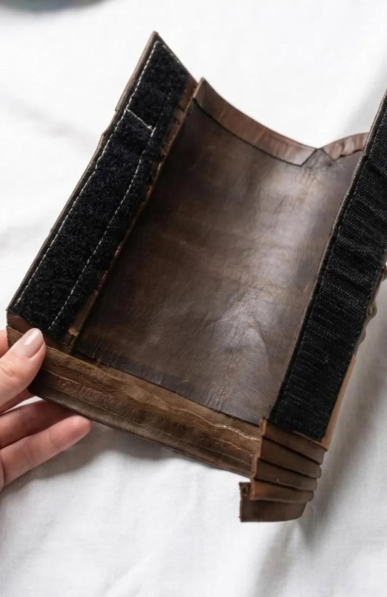 May include: A brown leather wrist cuff with black hook-and-loop fasteners. The cuff is made of layered leather and has a dark, aged appearance. The cuff is held by a hand against a white background.