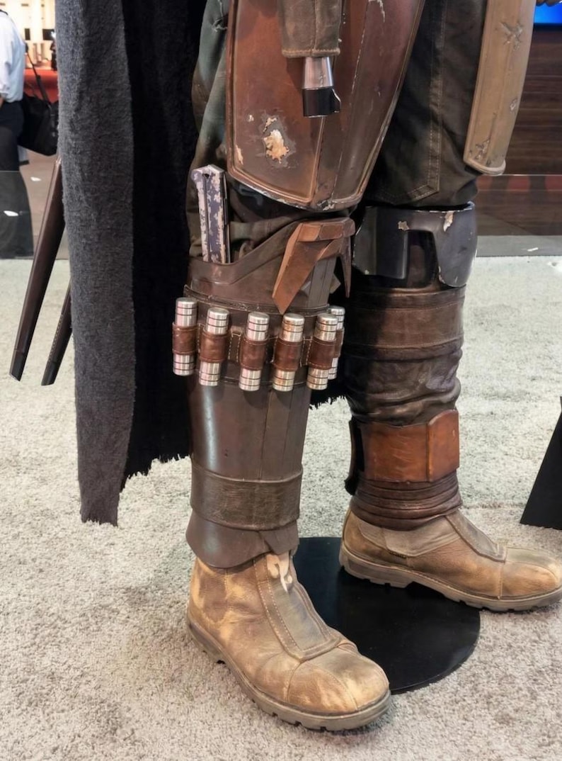 May include: Detailed view of a costume's lower leg armor, featuring brown leather boots and leg coverings. The armor includes a series of cylindrical metallic cartridges and a holster for a weapon. The costume has a weathered, battle-worn appearance.