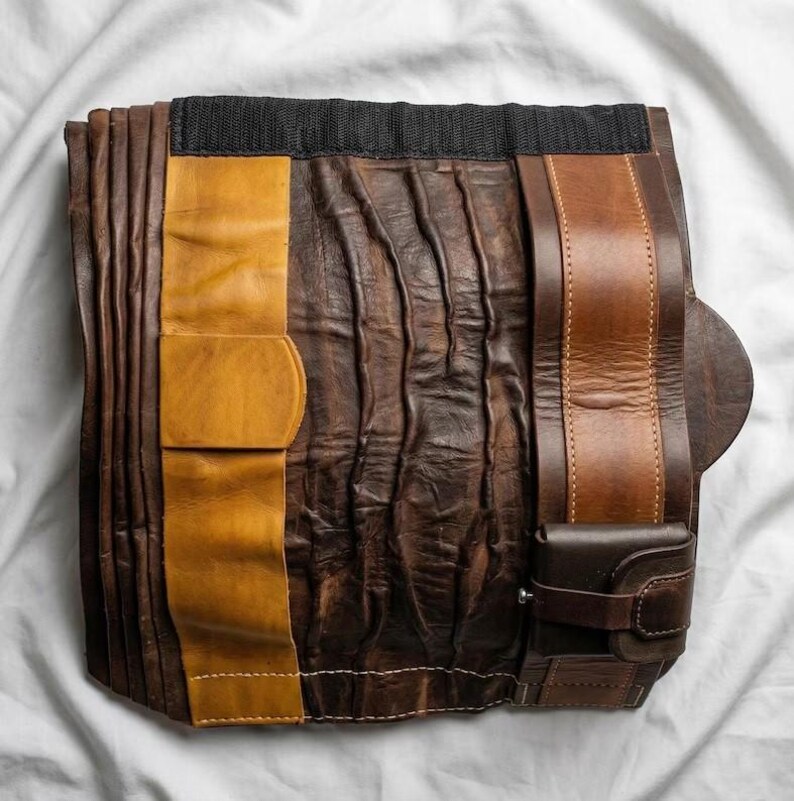May include: A handcrafted leather tool roll in shades of brown and tan, with a black elastic band at the top. The roll features multiple pockets and straps for securing tools. The leather has a textured, worn appearance.