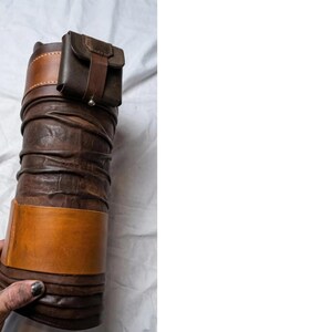 May include: A cylindrical, brown leather quiver with a small, attached pouch. The quiver has a light brown band around the middle and a darker brown band at the top. The pouch is secured with a strap and button.