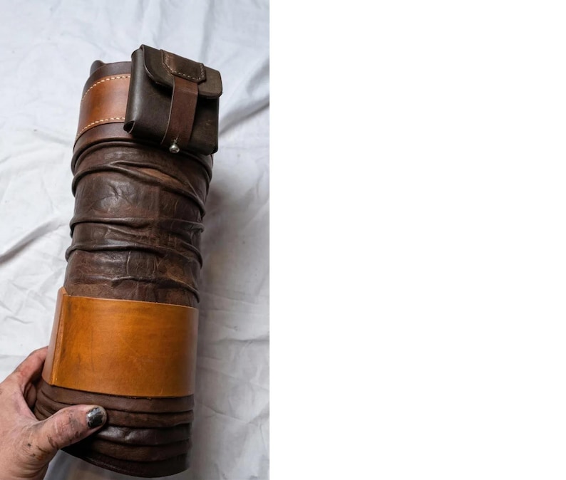 May include: A cylindrical, brown leather quiver with a small, attached pouch. The quiver has a light brown band around the middle and a darker brown band at the top. The pouch is secured with a strap and button.