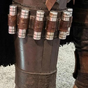 May include: Close-up of a brown leather leg armor with silver cylindrical details. The armor features a series of silver cylinders held in place by brown leather straps. The lower portion of the leg is covered in a brown leather boot.