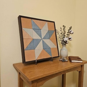 May include: A square wooden wall art piece featuring a geometric star pattern in shades of light blue, white, and natural wood tones, enclosed in a dark brown frame. The artwork is displayed on a small metal stand on a wooden table.