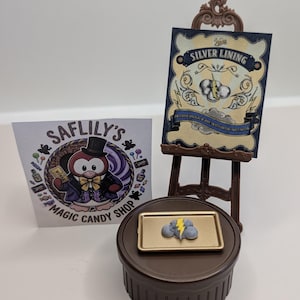 May include: A miniature brown easel displays a rectangular card titled Silver Lining with a cloud and lightning bolt graphic. A small gold tray holds a grey cloud-shaped item with a yellow lightning bolt. A square card shows a penguin logo.