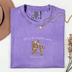 May include: A lilac Comfort Colors t-shirt featuring a monkey family on a scooter, the text "off to do kingdom work" and "Proverbs 31: 25-27". A gold star pendant necklace and a brown hat are also in the image.