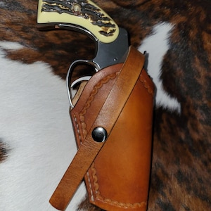 Könnte beinhalten: Ein Revolver mit einem strukturierten, cremefarbenen Griff mit dunkelbraunem, astähnlichem Muster, untergebracht in einem hellbraunen Lederholster. Das Holster hat einen gewellten Rand, sichtbare weiße Nähte und einen silberfarbenen Druckknopf.