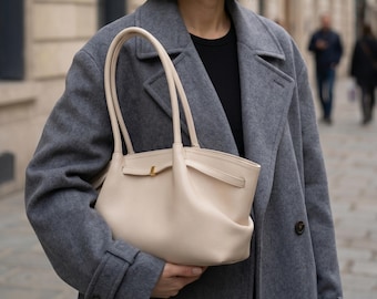 May include: A cream-colored leather tote bag with a structured design. The bag features a top handle, a gold-colored clasp, and a front pocket. The person is wearing a gray coat and a black shirt.