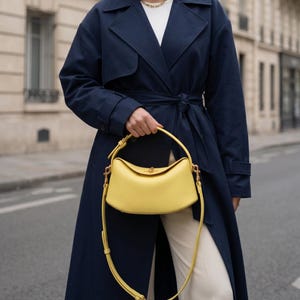 Könnte beinhalten: Eine leuchtend gelbe Handtasche mit geschwungenem Design, mit einem oberen Griff und einem langen Schulterriemen. Die Tasche wird vor einer Person gehalten, die einen marineblauen Trenchcoat und cremefarbene Hosen trägt. Eine goldene Halskette ist sichtbar.